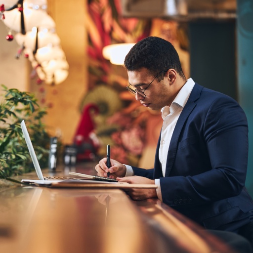 Side view of a focused young elegant man in eyeglasses taking notes in his notebook